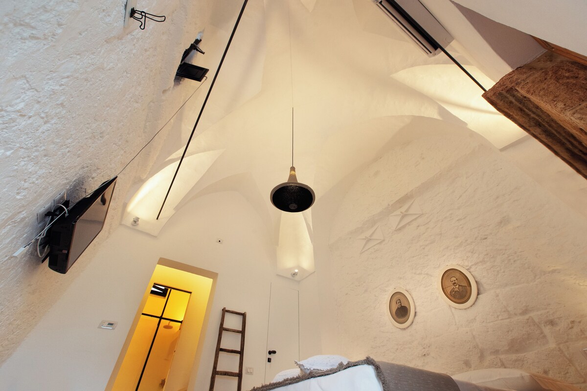 A high-ceiling room with a hanging light fixture, a ladder, and a door leading to another area.