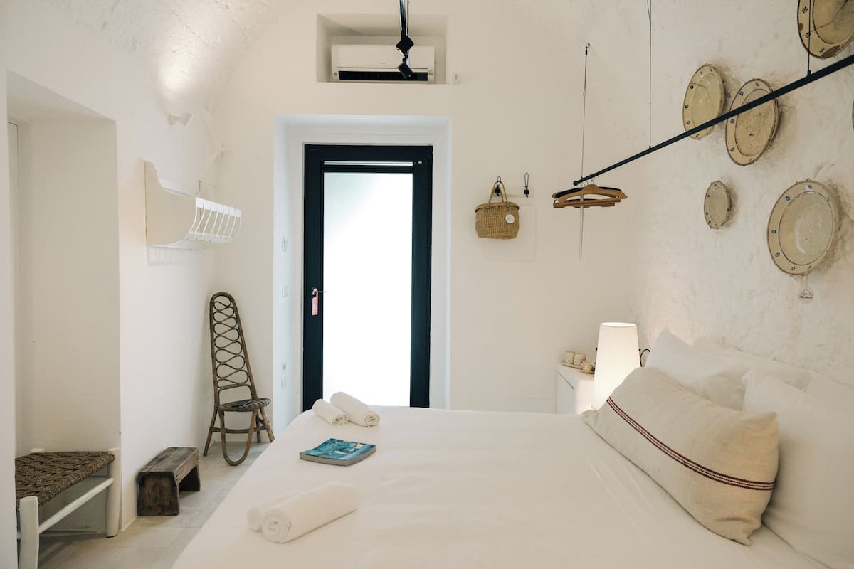 Quarto com cama, cadeira, mesa de cabeceira, abajur e porta.