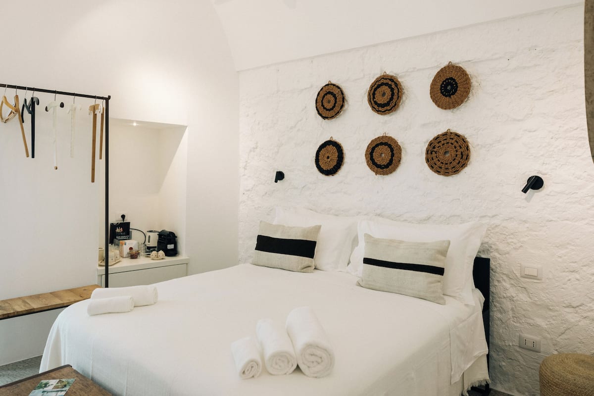 Uma cama bem arrumada com toalhas dobradas, uma mesa de cabeceira com cafeteira e um painel de parede com tapeçarias.