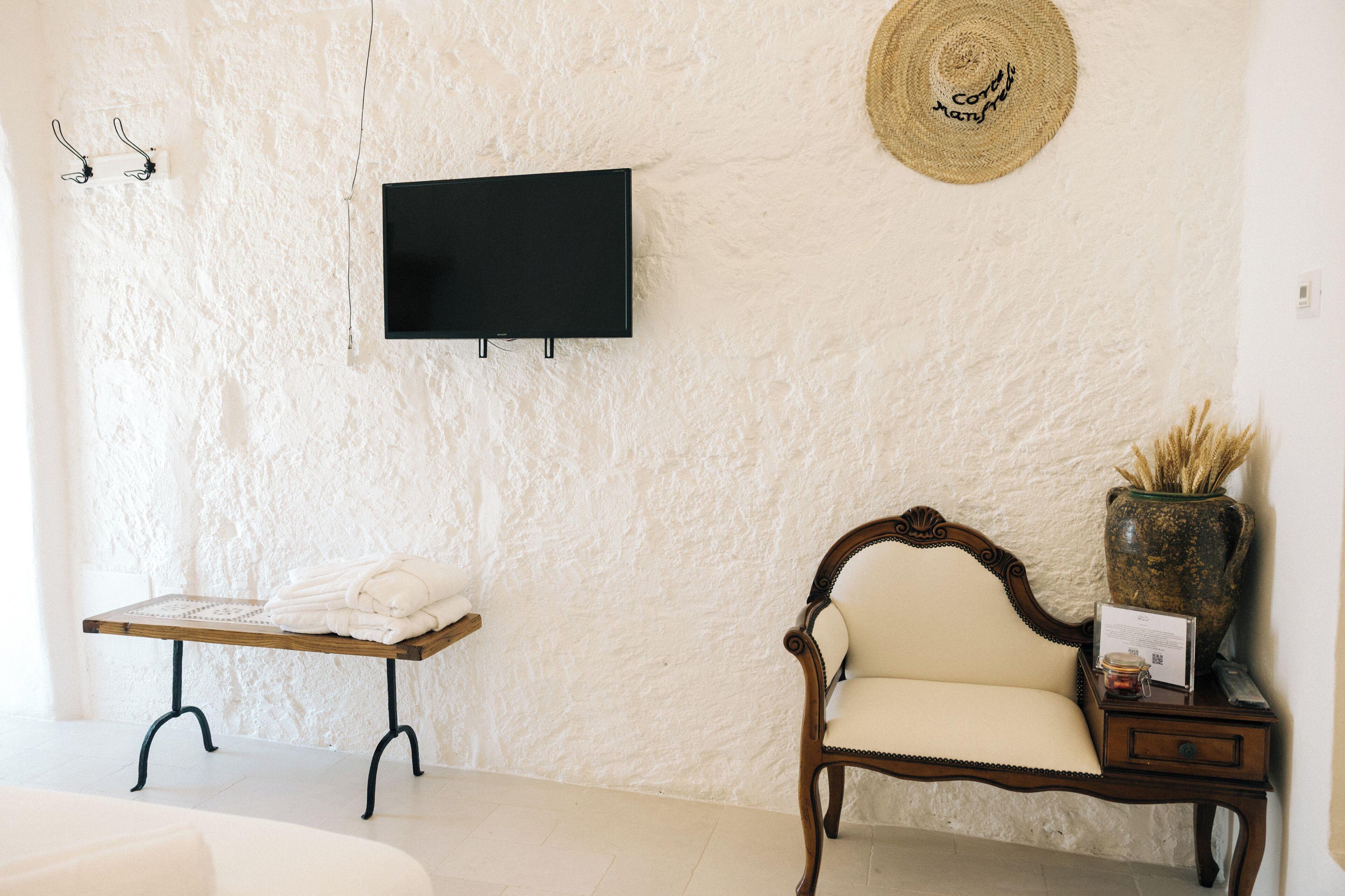 Una habitación con una televisión instalada en la pared, un banco de madera con una toalla, una silla acolchada y una mesita con un florero y un letrero.