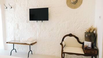 Una habitación con una televisión instalada en la pared, un banco de madera con una toalla, una silla acolchada y una mesita con un florero y un letrero.