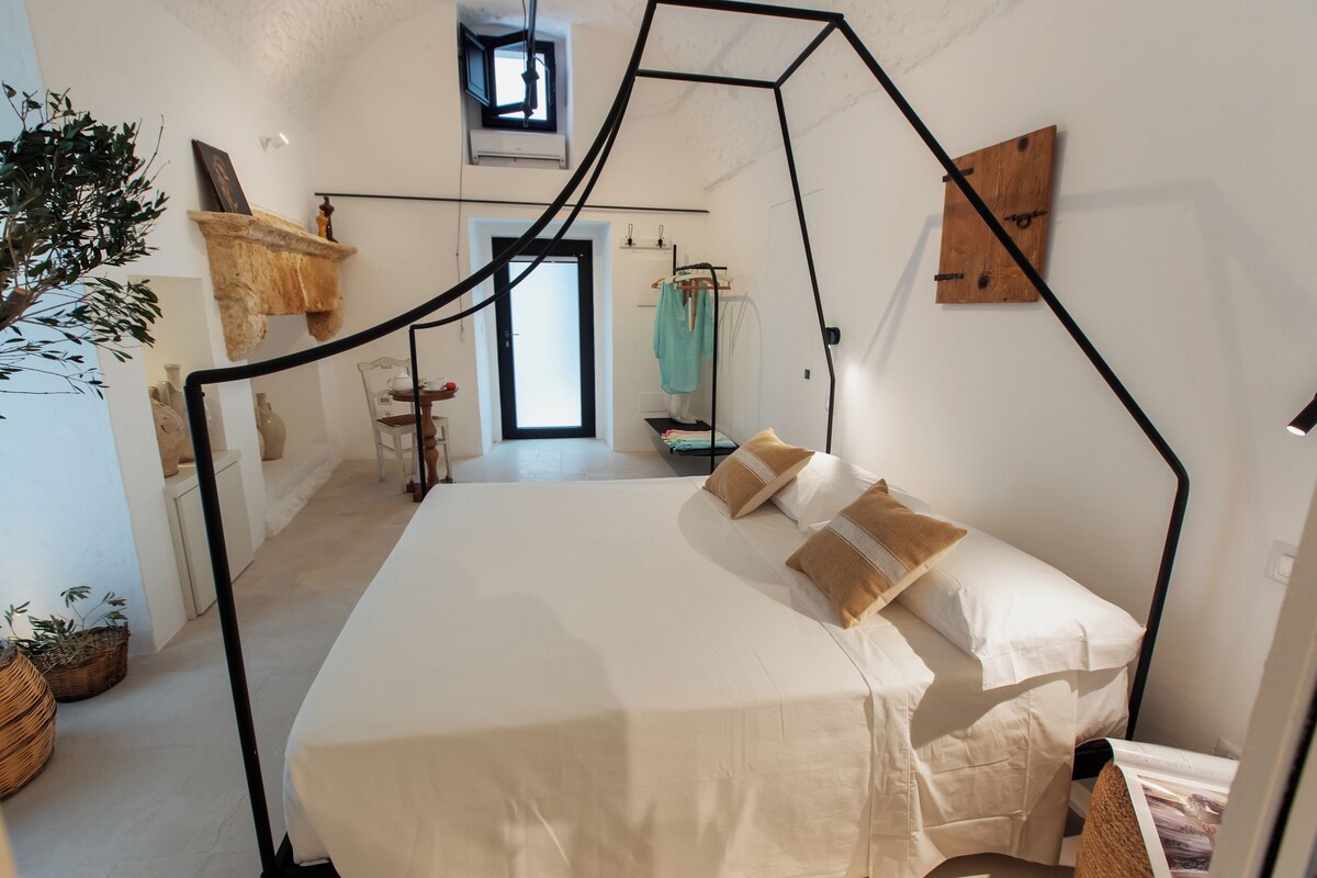 A bedroom with a bed, a hanging chair, a small table, a plant, and a window.