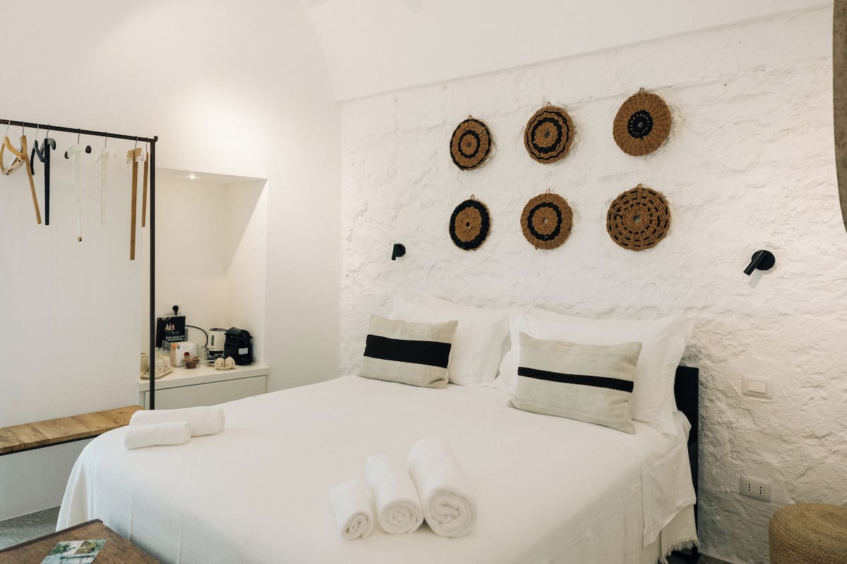 Uma cama bem arrumada com toalhas dobradas, uma mesa de cabeceira com cafeteira e um painel de parede com tapeçarias.