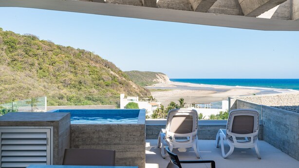 A balcony with two white chairs, a swimming pool, and a view of the beach and ocean. - Surf-Friendly Penthouse – Ocean Views, 3-Min Walk to the Beach (Bahias de Huatulco)