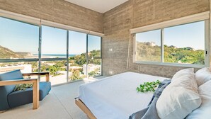 A bedroom with a large bed, a seating area with a chair, and a view of the ocean and hills. - Surf-Friendly Penthouse – Ocean Views, 3-Min Walk to the Beach (Bahias de Huatulco)