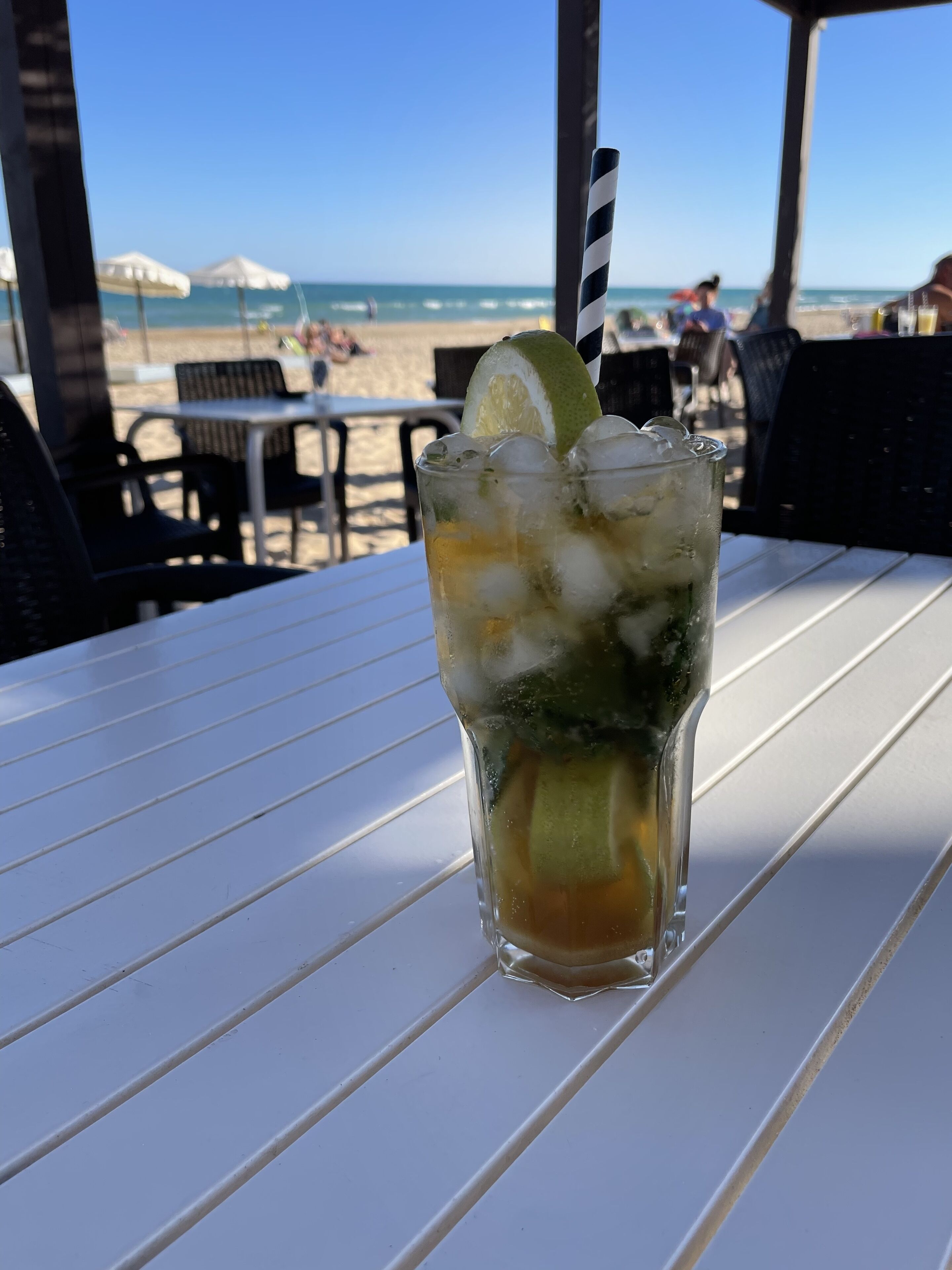 A glass of iced drink with a lime slice and a striped straw on a white table with a view of the beach and ocean in the background.