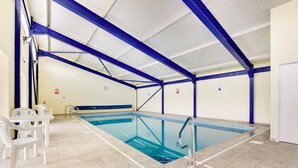 Une piscine intérieure avec des poutres bleues apparentes, un sol carrelé et des chaises en plastique blanches.