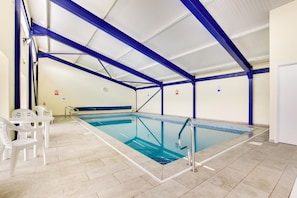 Une piscine intérieure avec des poutres bleues apparentes, un sol carrelé et des chaises en plastique blanches.
