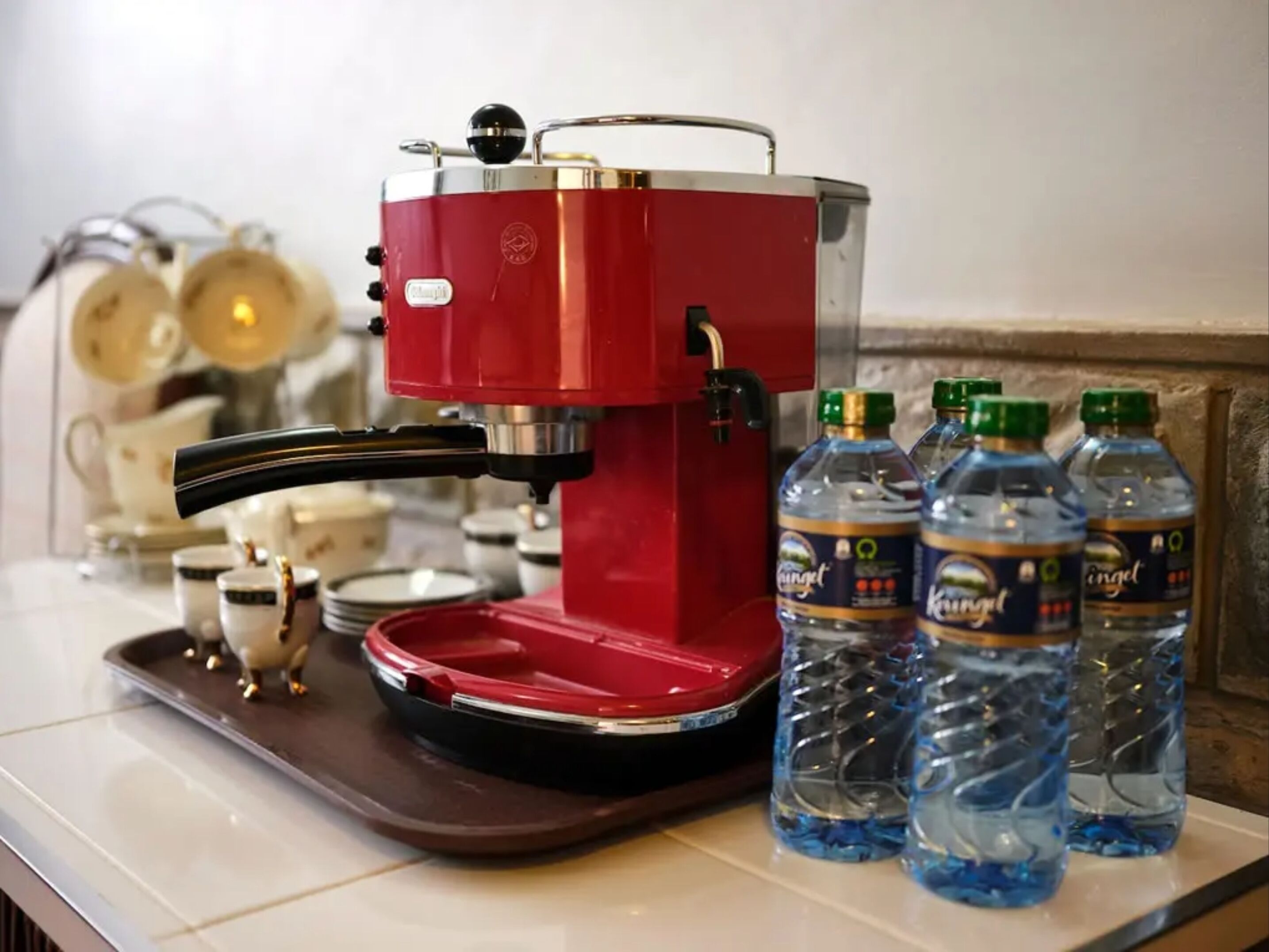 Une machine à café rouge, posée sur un plateau avec quatre bouteilles d’eau et quelques tasses.