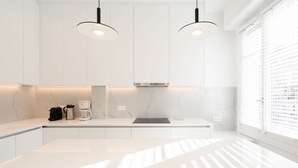 A modern kitchen with white cabinets, a stainless steel range hood, pendant lighting, and a window with blinds.