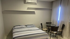 Un dormitorio con cama, una mesita con sillas, una ventana con cortinas y un equipo de aire acondicionado en la pared.