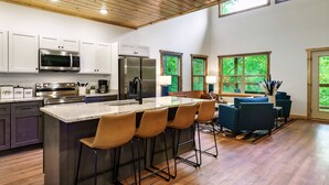 A modern kitchen with a central island, bar stools, a dining area with blue chairs, and large windows. - Smokies Private Pool Cabin, Sleeps 14,Theatre & Game Room, Minutes from Parkway! (Sevierville)