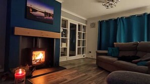 A cozy living room with a wood-burning stove, a sofa, and a large window with curtains. - Benllech Sea View bungalow, Dog Friendly sleeps 6 (Benllech)