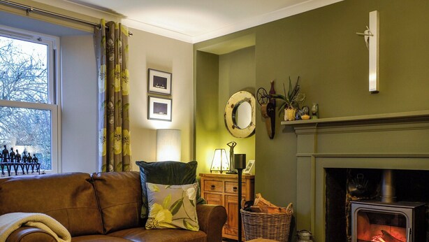 A cozy living room with a brown leather sofa, a wooden coffee table with books, a fireplace, and a window with curtains. - A real gem of a cottage. (Alyth, near Blairgowrie)