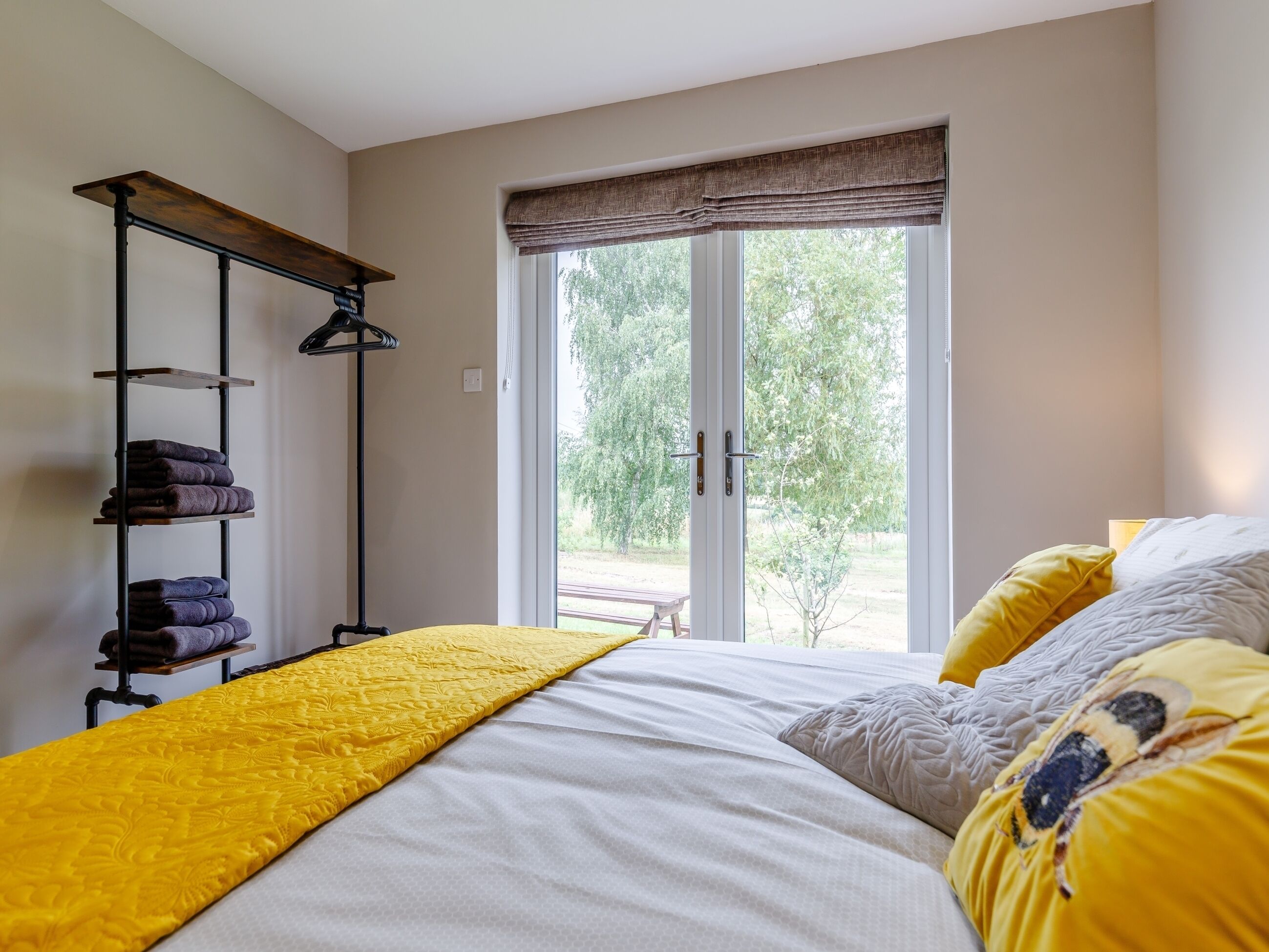 A bedroom with a bed, a window with a view of greenery, a shelf with towels, and a bedside table.