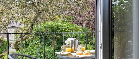 Un balcón con una mesa dispuesta para el desayuno, que incluye jugo de naranja y pastelería, rodeado de exuberante vegetación y un edificio de piedra al fondo.