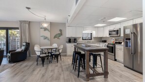 Una cocina moderna con armarios blancos, electrodomésticos de acero inoxidable y un comedor con mesa y sillas de madera.