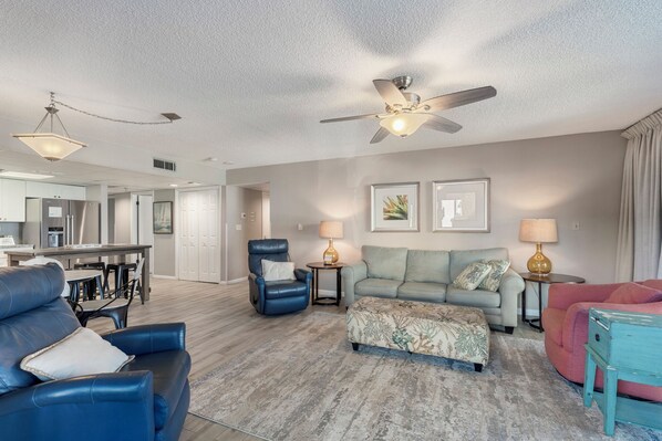 A spacious living room with a ceiling fan, a sofa, a chair, and a coffee table. There is a kitchen area in the background.