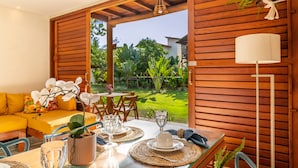 Un espacio de sala-comedor abierto con una mesa para dos personas, rodeado de muebles de madera y vistas a un jardín exuberante.