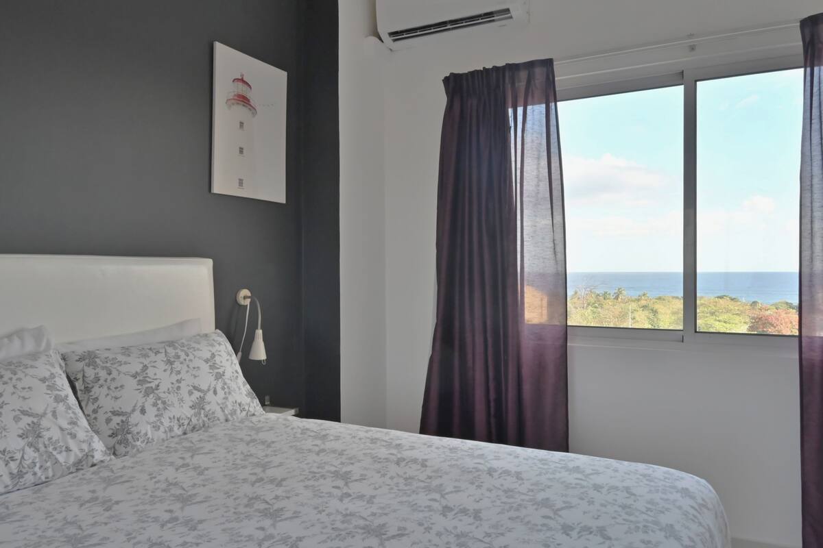 A bedroom with a bed, a nightstand, a wall-mounted air conditioner, a window with curtains, and a framed picture on the wall.