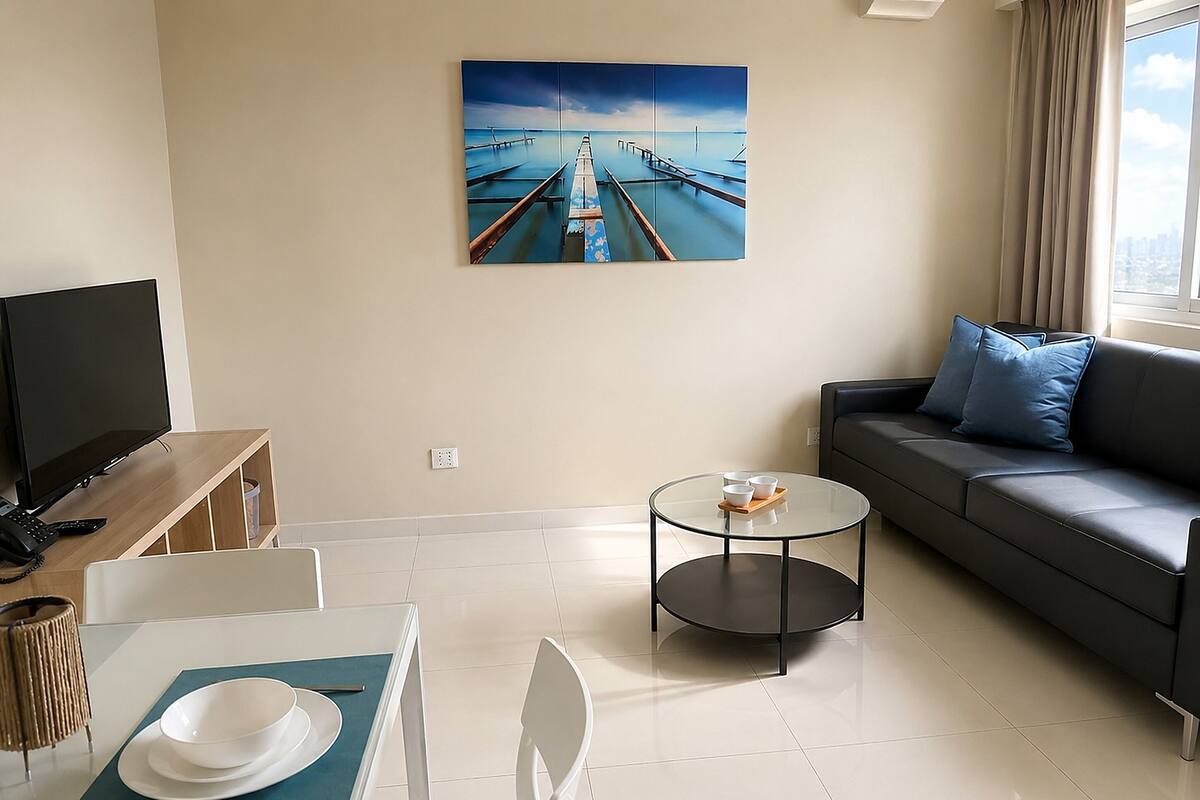 A modern living room with a flat-screen TV, a dining table with chairs, a glass coffee table, a sofa with cushions, and a painting on the wall.