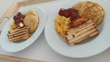 Duas travessas de café da manhã com panquecas, ovos mexidos, linguiça e pão tostado em uma bandeja.