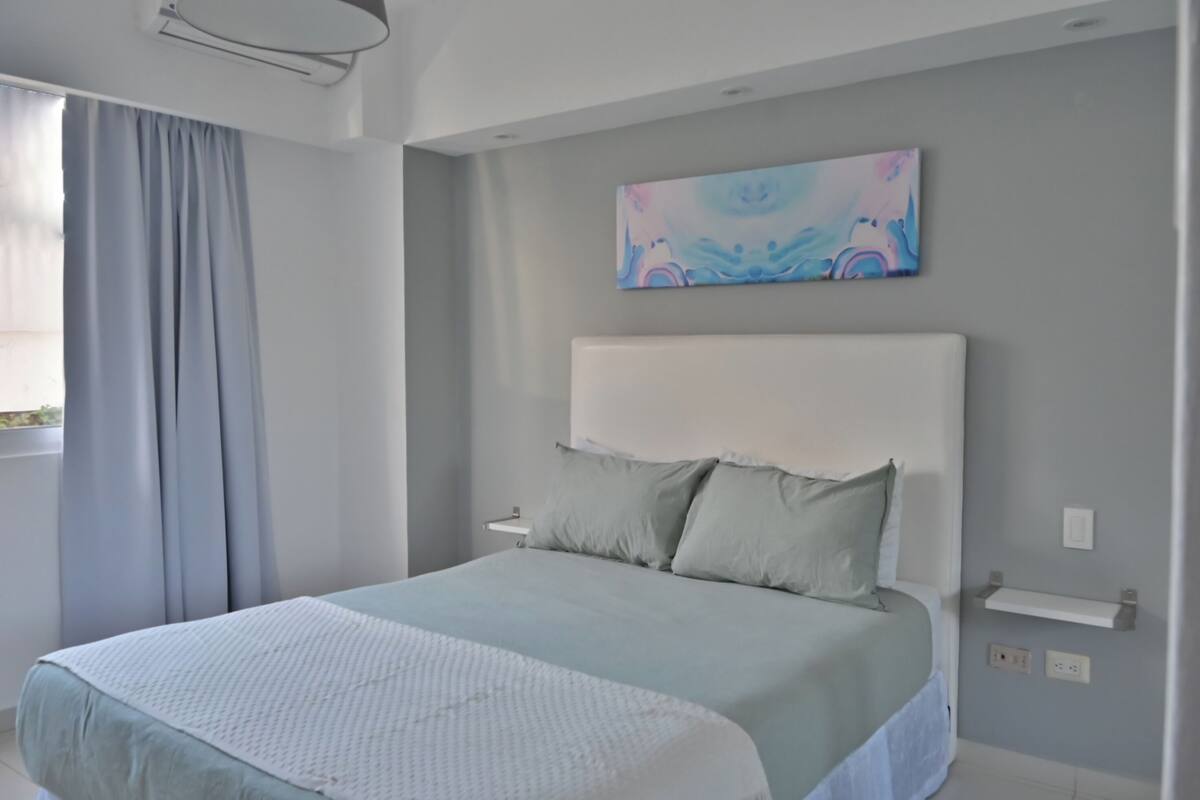 A modern bedroom with a large bed, a headboard, a painting, and a window with curtains.