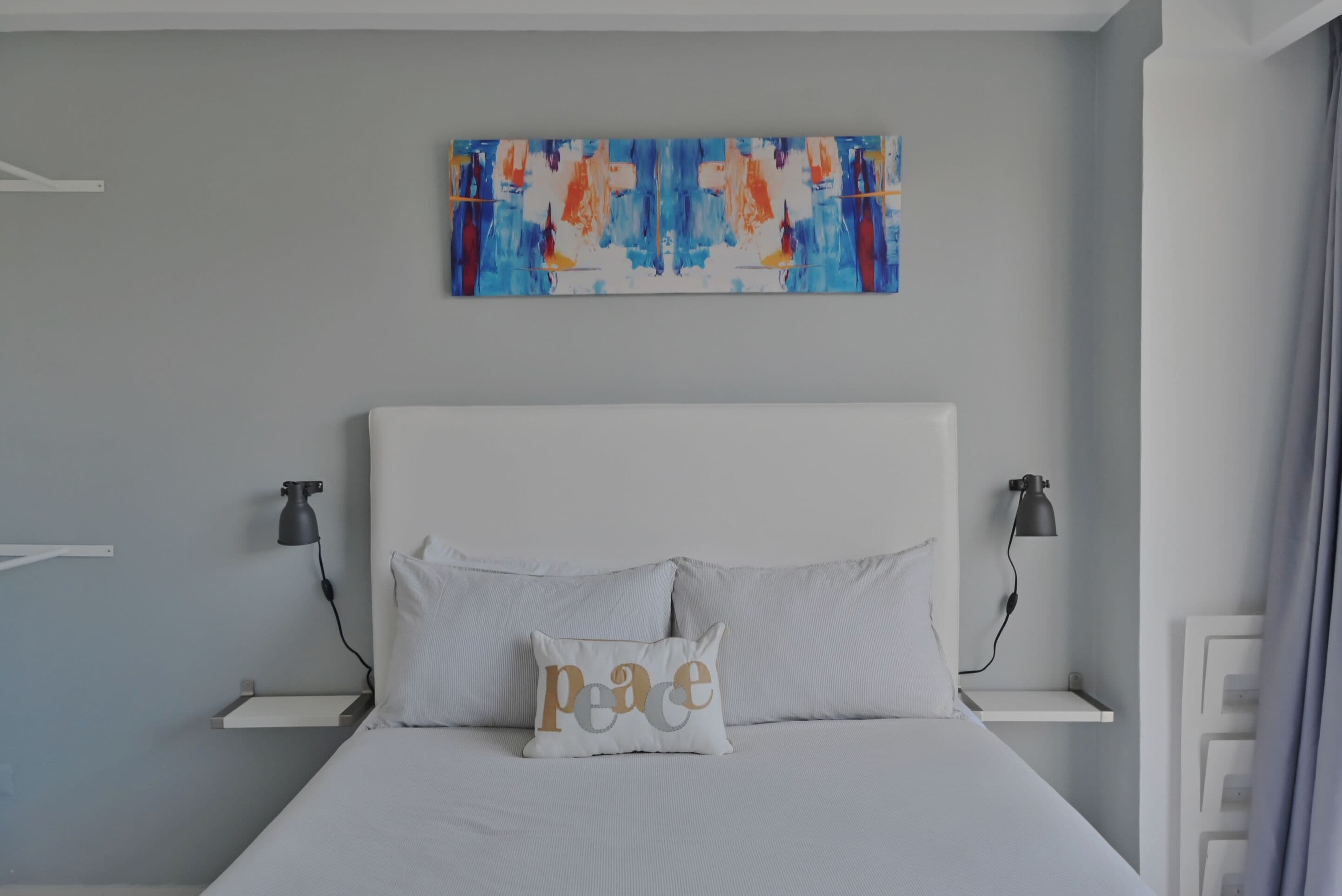Quarto com cama branca, duas mesas de cabeceira com luminárias, uma obra de arte na parede e um travesseiro com a palavra 'peace'.