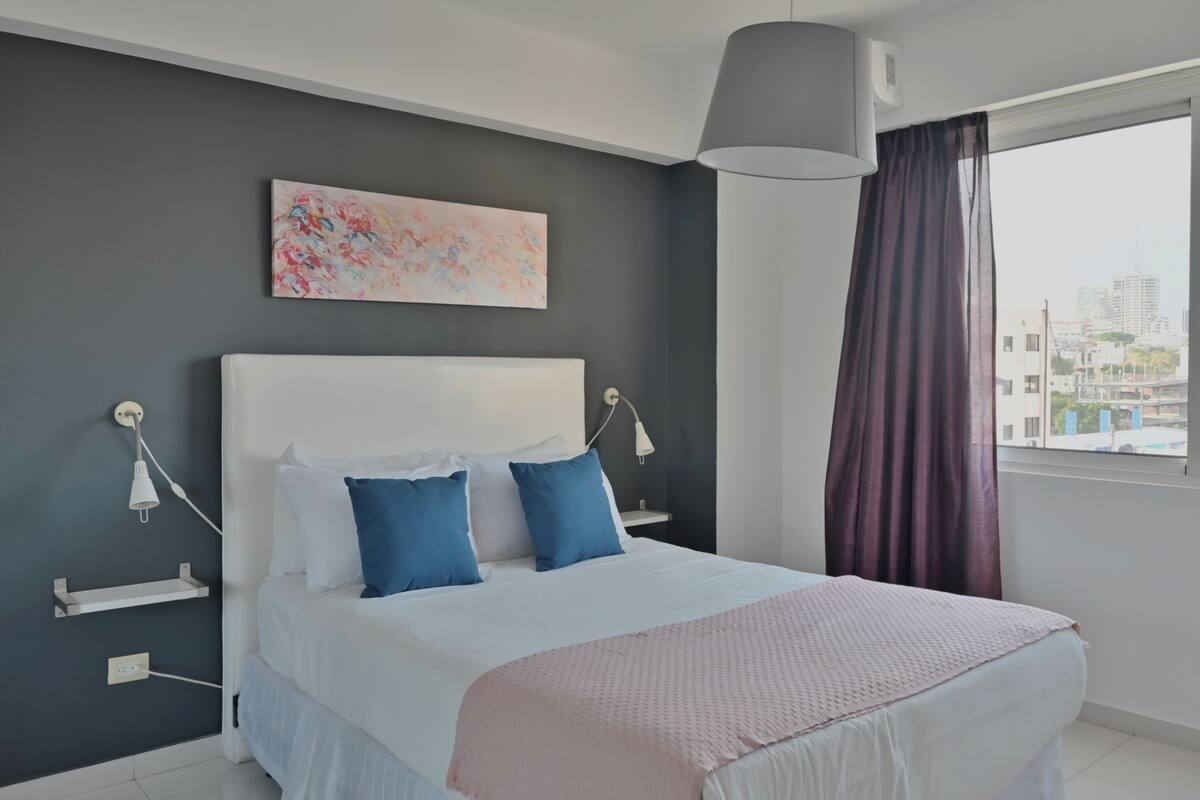 A modern bedroom with a large bed, a city view, and a gray ceiling.