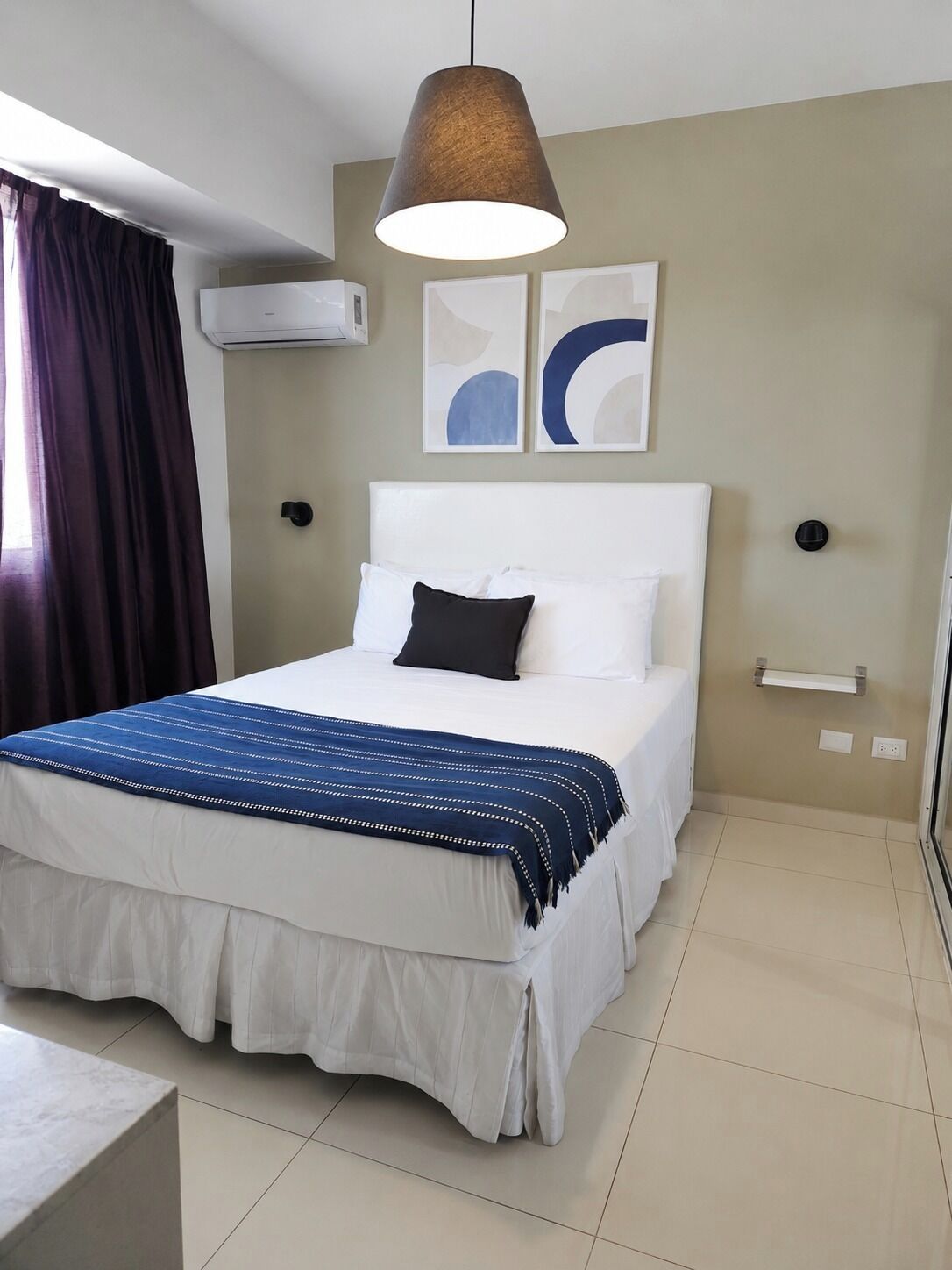 Habitación de hotel con cama, lámpara colgante, equipo de aire acondicionado y arte abstracto en la pared.