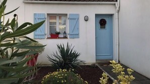 A white building with a blue door and shutters, a small window with a red planter, and a garden with plants and flowers. - House in a quiet, sought-after area (Saint-Gilles-Croix-de-Vie)