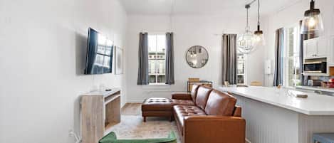 Een moderne woonkamer met een bruine leren bank, een groene poef, een flatscreen-tv en een keukenhoek met een wit aanrecht en hangende pendelverlichting.