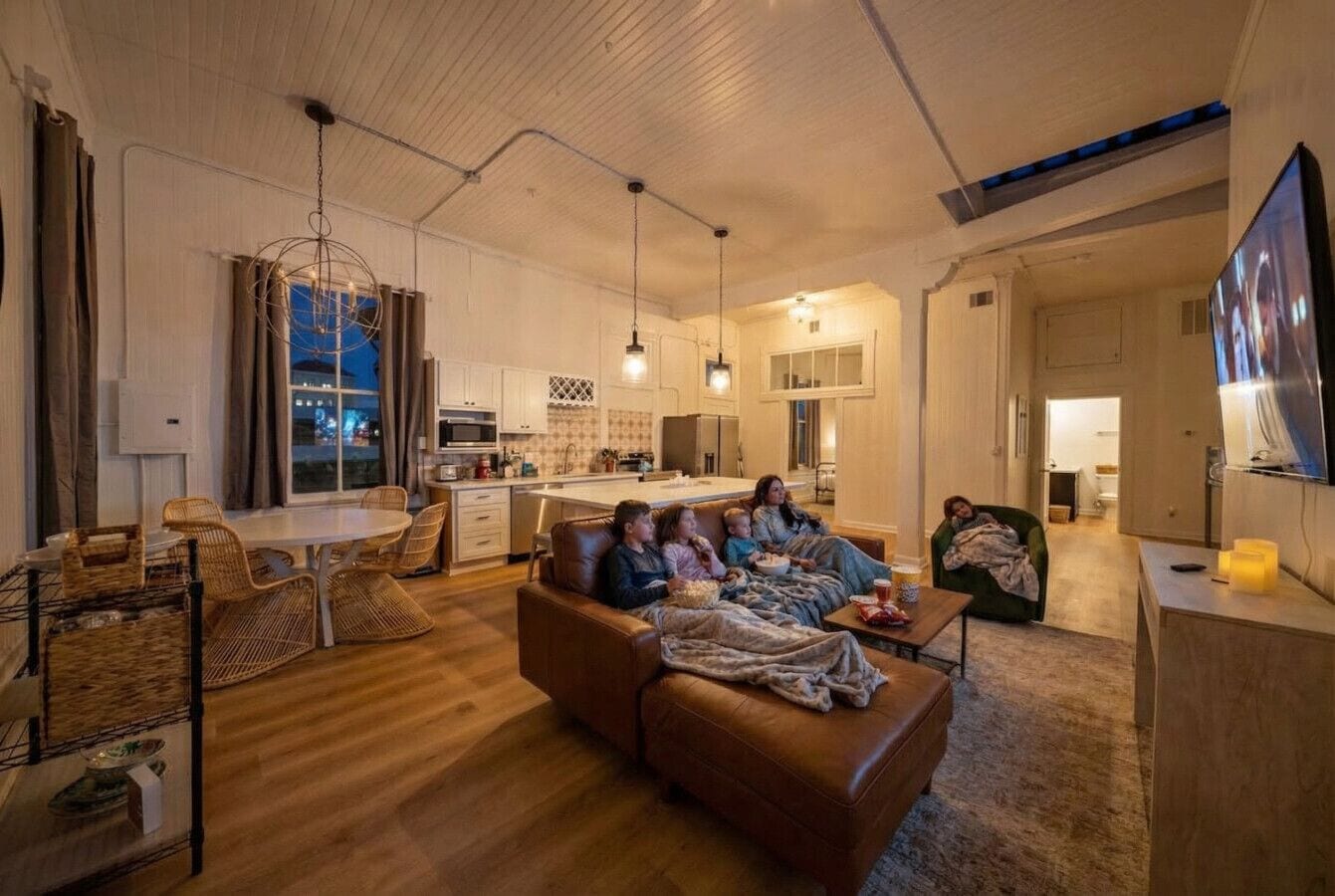 A cozy living room with a brown leather sofa, a dining table with wicker chairs, and a kitchen in the background.