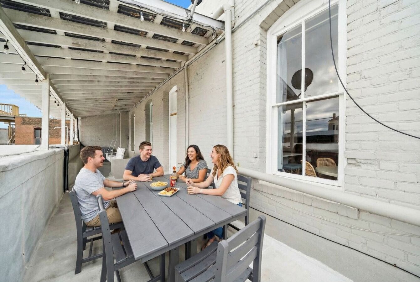 Un patio techado con una mesa y sillas, un muro de ladrillo blanco y una ventana con sillones de mimbre en el interior.