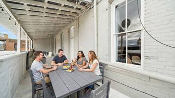 Un patio techado con una mesa y sillas, un muro de ladrillo blanco y una ventana con sillones de mimbre en el interior.
