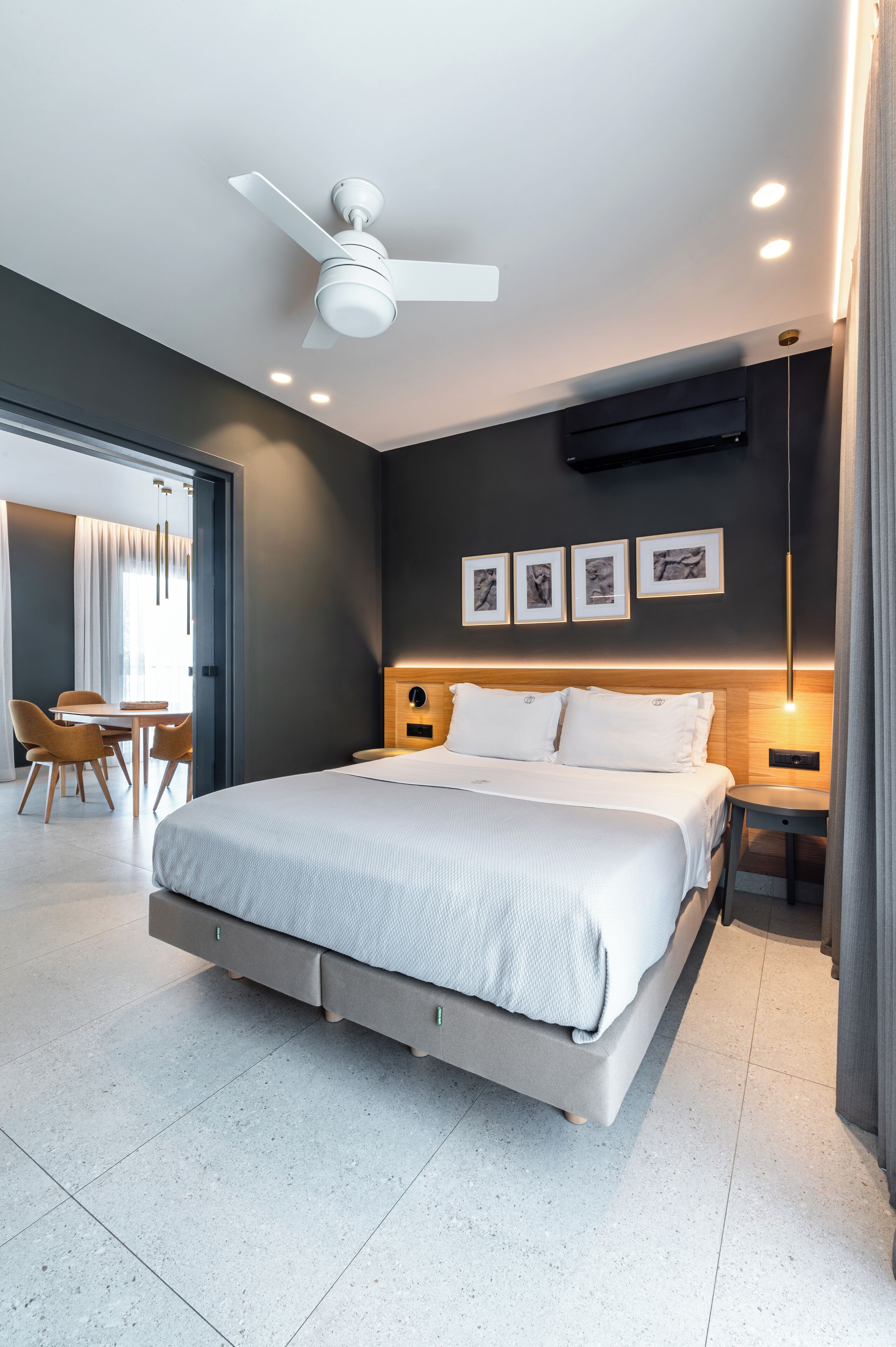 Una habitación de hotel moderna con una cama grande, ventilador de techo, iluminación empotrada, un área de comedor con sillas e imágenes enmarcadas en la pared.