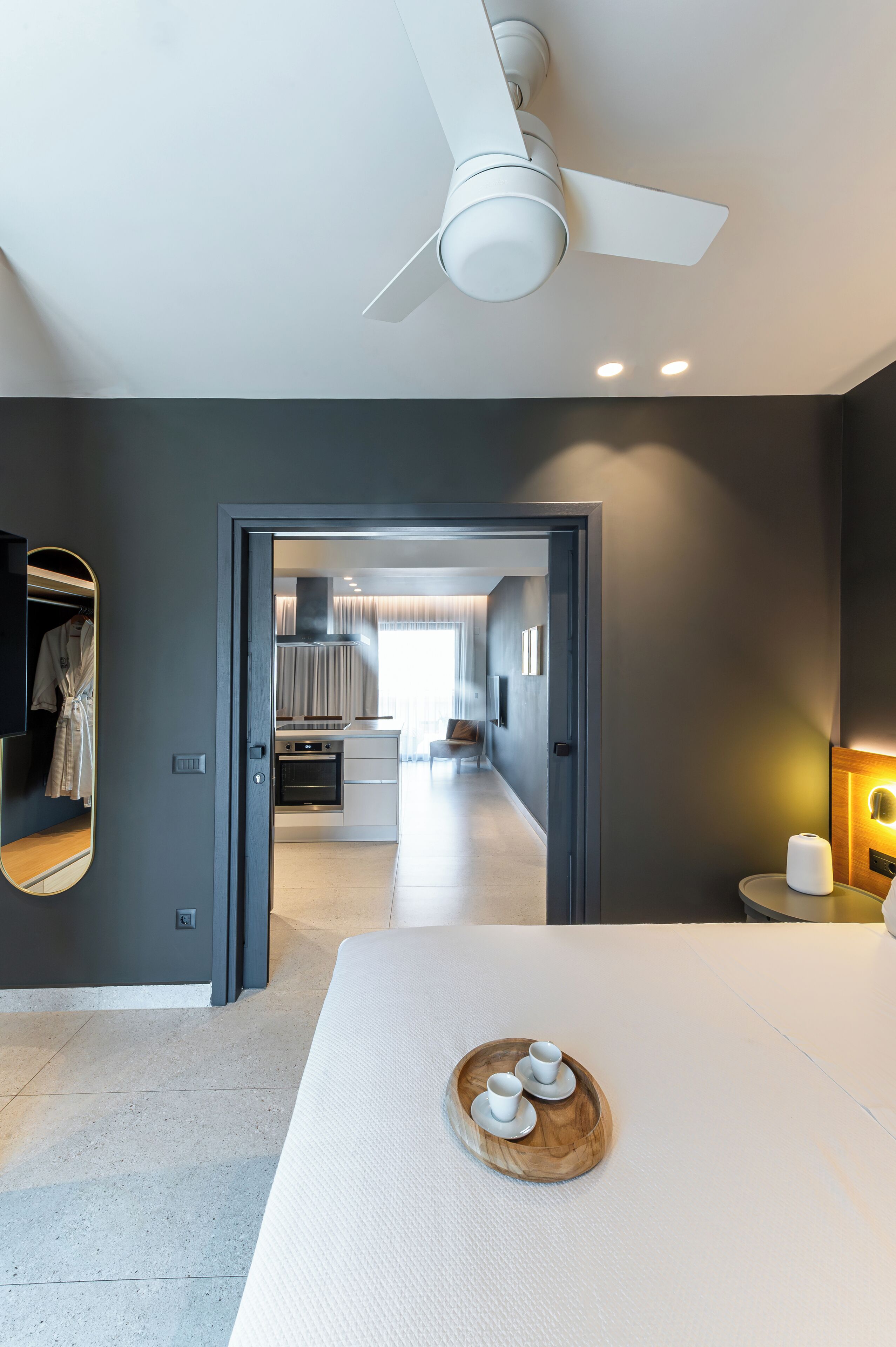 Una moderna camera d'hotel con un letto grande, una ventola a soffitto, un tavolo da pranzo con un vassoio di tazze, uno specchio e vista sulla cucina e sull'area soggiorno.