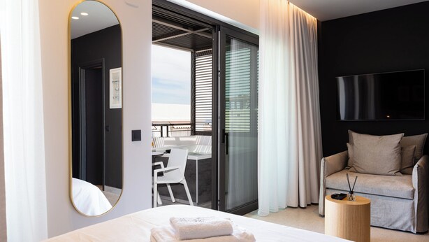 A modern hotel room with a large bed, a round wooden side table, a sofa, a TV mounted on the wall, and a sliding glass door leading to a balcony. - Porta Suites (Heraklion)