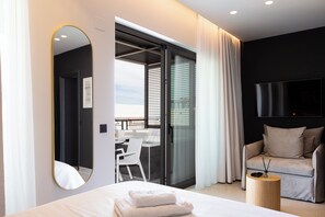 A modern hotel room with a large bed, a round wooden side table, a sofa, a TV mounted on the wall, and a sliding glass door leading to a balcony. - Porta Suites (Heraklion)