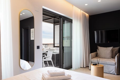 A modern hotel room with a large bed, a round wooden side table, a sofa, a TV mounted on the wall, and a sliding glass door leading to a balcony.