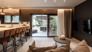 A modern living room with a sofa, a coffee table, and a TV. There is a kitchen area with a bar and chairs. - Porta Suites (Heraklion)