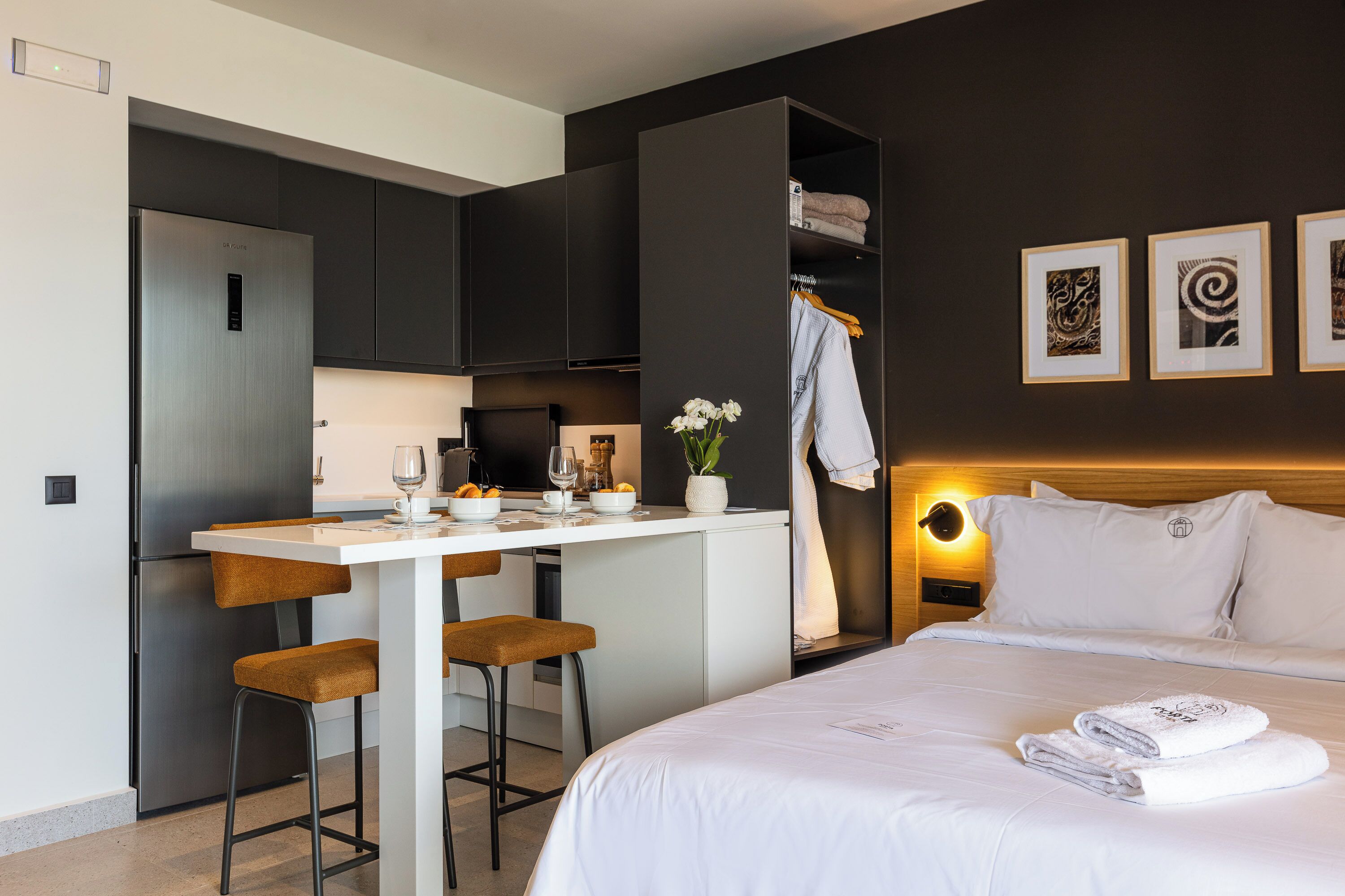 A modern hotel room with a bed, a desk with two chairs, a small kitchen area, and artwork on the wall.