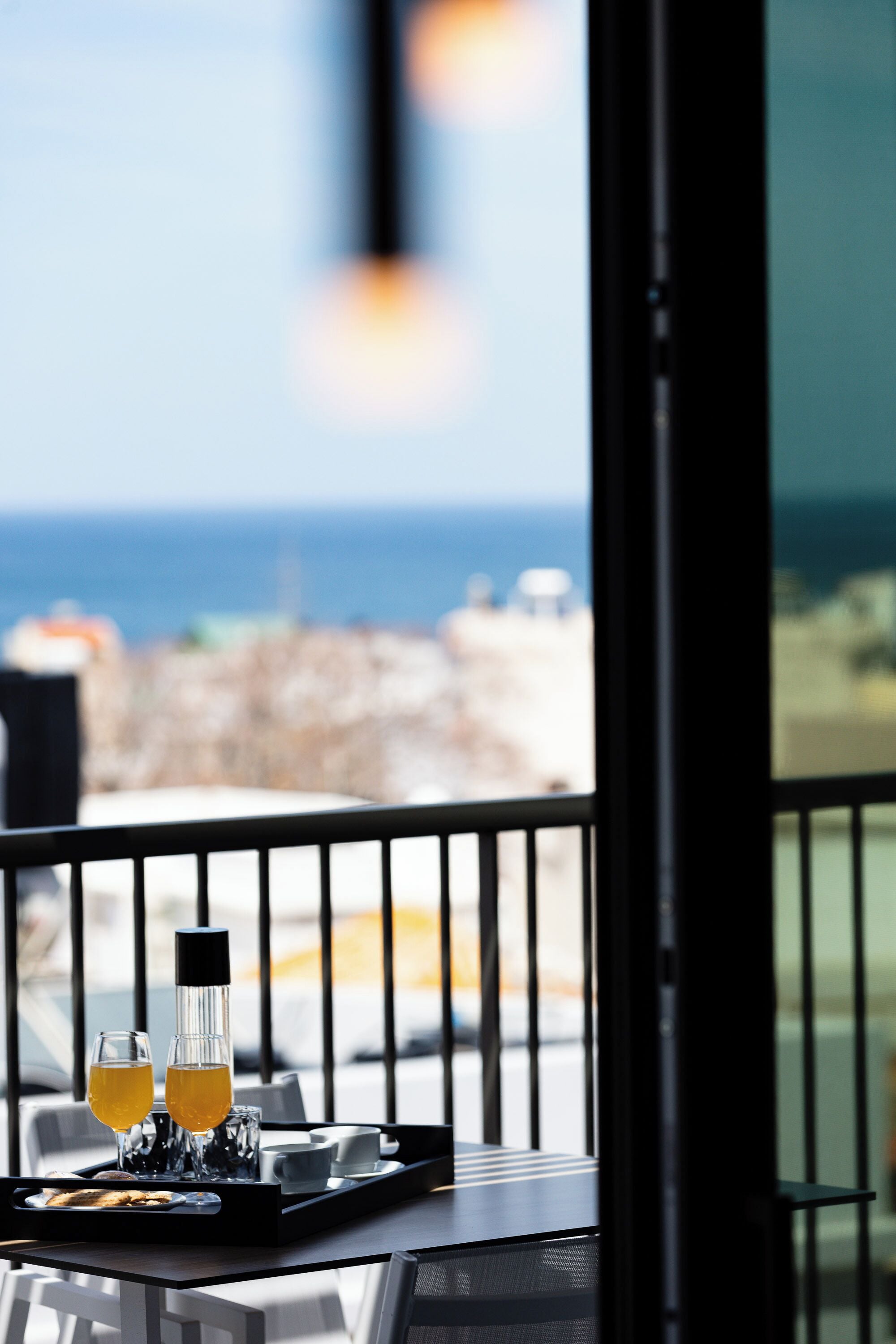 Un tavolo con un vassoio contenente due bicchieri di succo d'arancia, una bottiglia e un piatto con cibo. Il tavolo si trova vicino a una finestra che offre vista sul mare e sugli edifici.