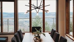 Ein Essbereich mit einem langen Holztisch, schwarzen Stühlen und einer modernen Pendelleuchte. Große Fenster bieten Blick ins Grüne.