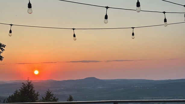 Eine Terrasse im Freien mit Holzmöbeln und Lichterketten, mit Blick auf eine reizvolle Landschaft bei Sonnenuntergang.