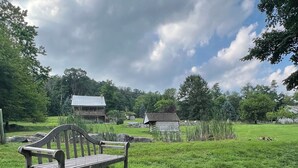 A rustic wooden bench in a grassy field with a moss-covered rock nearby, a wooden cabin in the background, and a cloudy sky.