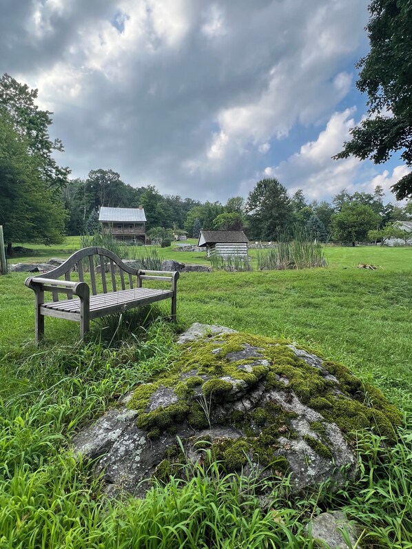 A rustic wooden bench in a grassy field with a moss-covered rock nearby, a wooden cabin in the background, and a cloudy sky.