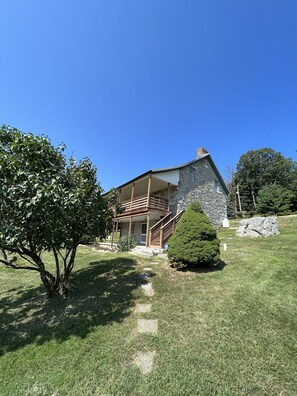 A stone house with a wooden balcony, surrounded by a grassy yard and a large tree.