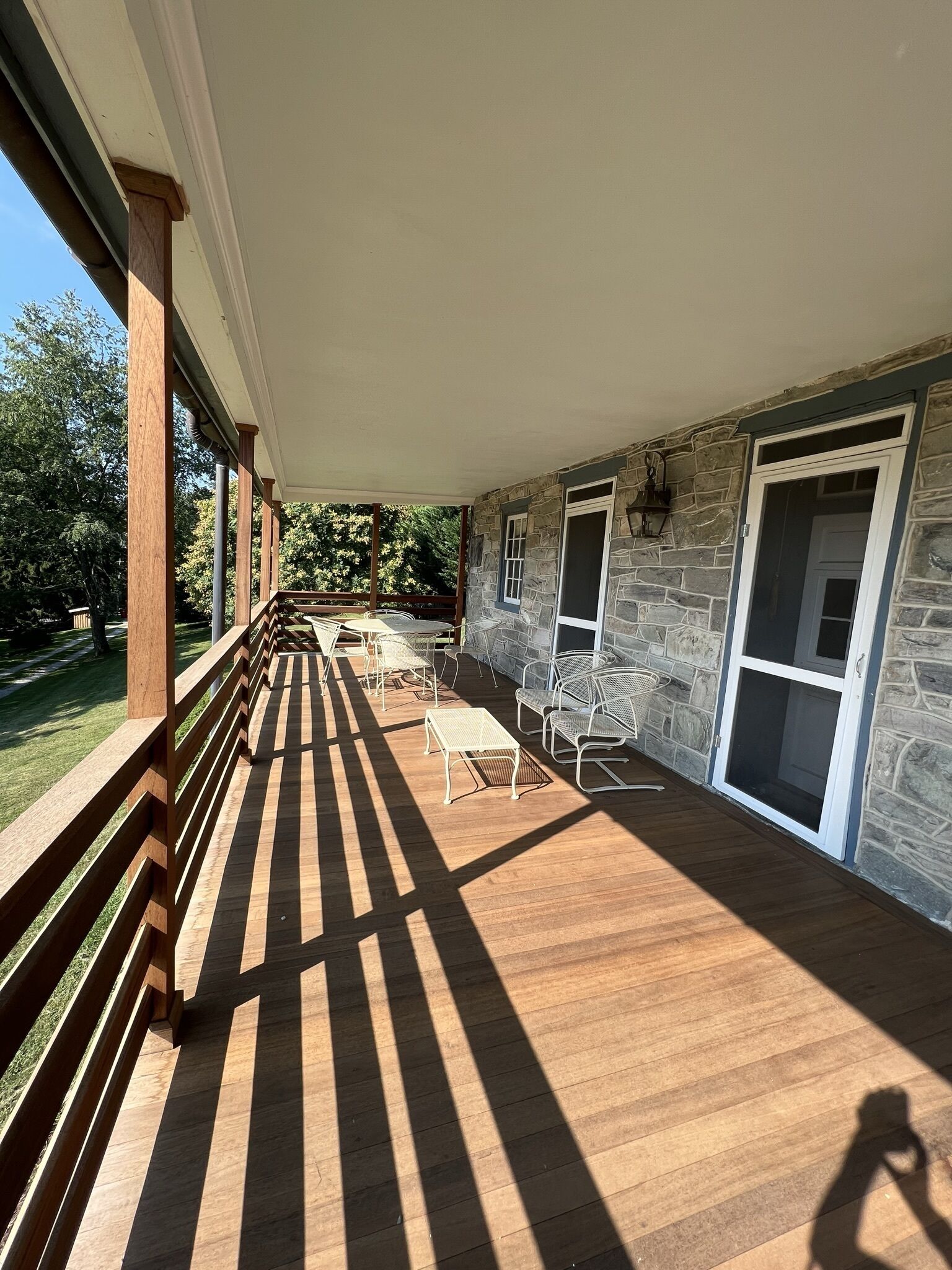 A covered outdoor patio with wooden flooring, stone walls, and a view of the surrounding greenery.