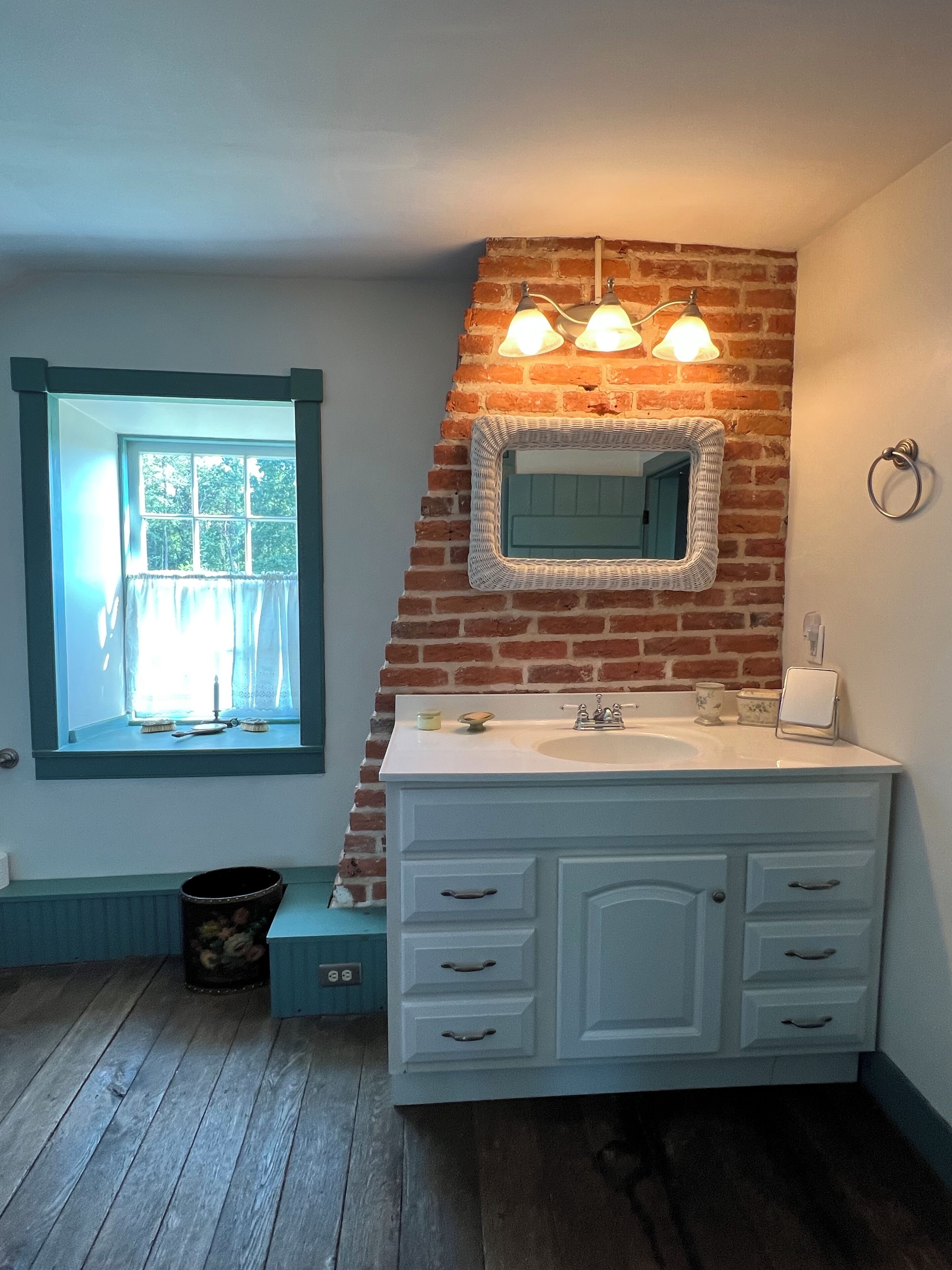 A bathroom with a white vanity, a mirror with a wicker frame, and a window with blue trim.